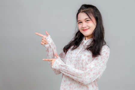 Young Asian woman smiling brightly while pointing sideways with both hands, showing a cheerful and inviting expression. Ideal for promotional ads, presentations, or marketing concepts.の写真素材