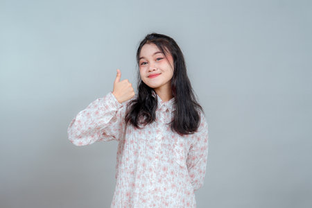 Cheerful young Asian woman showing thumbs up with a confident smile, expressing positivity and approval. Studio portrait with casual floral outfit and neutral background.の写真素材