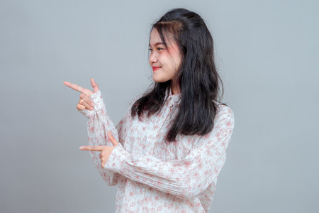 Side view of a smiling young Asian woman pointing sideways with both hands. Her cheerful and playful expression is perfect for use in advertising, promotion, or creative concepts.の写真素材