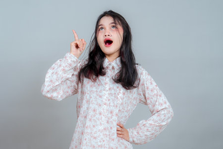 Surprised Asian woman with wide open mouth and raised finger, expressing sudden realization or brilliant idea. Studio portrait on gray background, emotional reaction.の写真素材