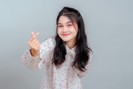 Smiling young Asian woman showing finger heart gesture with a soft, sincere expression. Portrait against gray background, ideal for friendly, cheerful, and emotional advertising themes.の写真素材