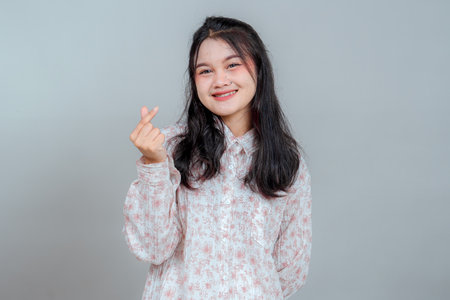 Young Asian woman smiling warmly while making a finger heart gesture, showing a cheerful and affectionate expression, perfect for friendly, love, or lifestyle concepts.の写真素材
