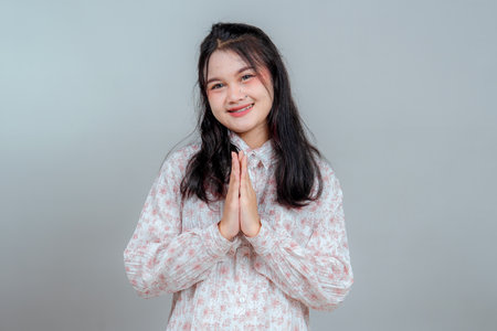 Smiling young Asian woman with long black hair making a respectful greeting gesture with both hands together. Her warm facial expression suits cultural, hospitality, and welcome-themed ads.の写真素材