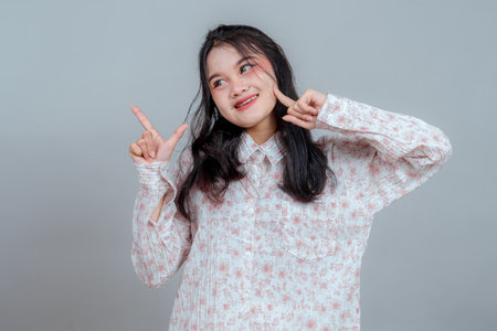 Cheerful young Asian woman smiling brightly while making playful finger gestures and looking sideways. Her expressive face and joyful pose exude positivity, charm, and energy.の写真素材