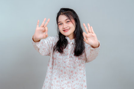 Smiling young Asian woman with long black hair showing double OK hand signs towards the camera. Her cheerful and confident expression makes her ideal for positive advertising themes.の写真素材