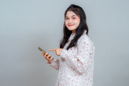 Young woman smiling sweetly while pointing at her smartphone, showing confident eye contact and positive facial expression, ideal for tech, lifestyle, or communication advertising.の写真素材