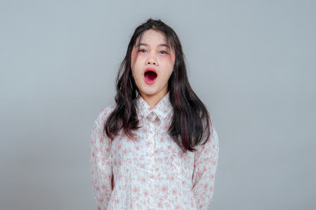 Young Asian woman with wide-open mouth and surprised expression, standing against a plain background. Her shocked face and body posture express disbelief, awe, or sudden realization.の写真素材