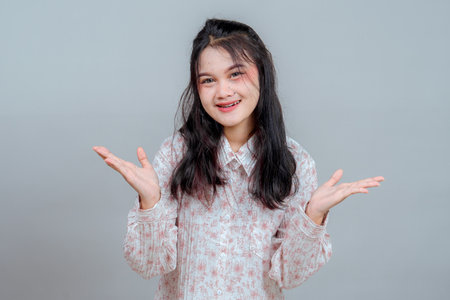 Portrait of a young Asian woman smiling with a confused expression, raising both hands. She wears a floral blouse and stands against a plain gray background.の写真素材