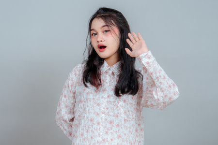 Curious young Asian woman raising her hand to ear, showing a listening gesture with a surprised facial expression. Studio portrait for advertising attention and curiosity concepts.の写真素材