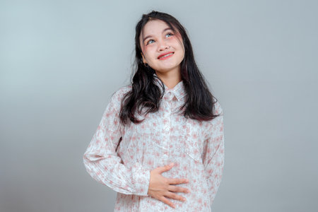 Portrait of a young Asian woman smiling gently with a dreamy expression, looking up while holding her stomach. She wears a floral blouse, standing against a neutral gray background.の写真素材