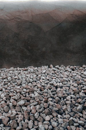 Minimalist textured background of natural stone gravel against a smooth, dark plastered wall. The composition offers contrast, raw material texture, and negative space for design flexibility.の写真素材