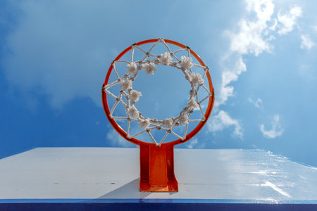 A low-angle shot of a basketball hoop against a bright blue sky. The vibrant orange ring and white...