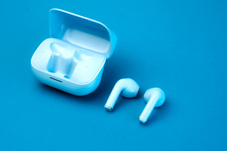 A pair of modern wireless earbuds placed next to an open charging case on a blue background. A sleek, stylish, and compact audio accessory perfect for daily use, music, and calls.の写真素材