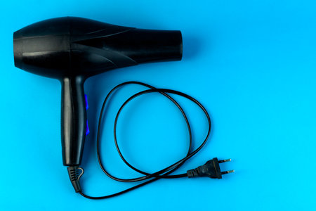 A black electric hair dryer with a long power cord on a blue background. A high-powered blow dryer...