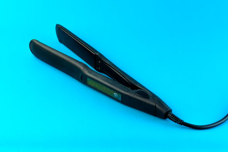 A black electric hair straightener with a digital display and a long power cord on a blue background. A professional flat iron designed for smooth and sleek hair styling with precision.の写真素材