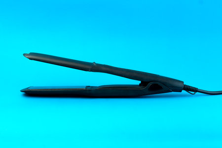 A black electric hair straightener with a digital display and a long power cord on a blue background. A professional flat iron designed for smooth and sleek hair styling with precision.の写真素材