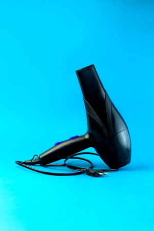 A black electric hair dryer with a long power cord on a blue background. A high-powered blow dryer designed for fast drying and styling. Ideal for professional salons and home use.の写真素材