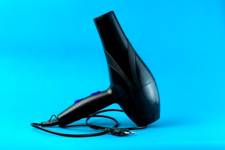 A black electric hair dryer with a long power cord on a blue background. A high-powered blow dryer designed for fast drying and styling. Ideal for professional salons and home use.の写真素材