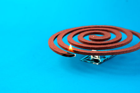 A burning mosquito coil on a metal stand against a blue background. Traditional insect repellent used to keep mosquitoes away, providing protection for indoor and outdoor spaces.の写真素材