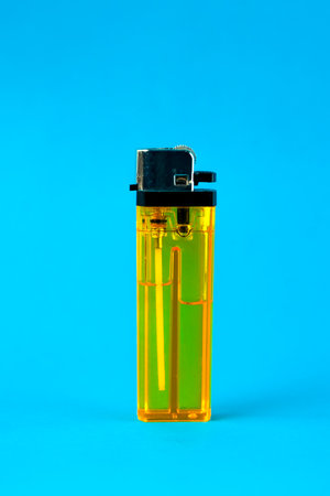 A transparent yellow plastic lighter standing upright against a bright blue background. The close-up shot highlights its metallic ignition mechanism and fluid inside, ideal for fire needs.の写真素材