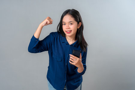 Young Asian woman smiling confidently while flexing her arm and holding a smartphone, showing a...