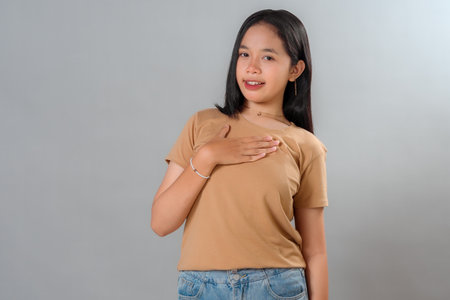 A young Asian woman with a sincere facial expression, placing hand on her chest and smiling slightly, conveying gratitude, honesty, or a heartfelt emotion in a casual pose.の写真素材