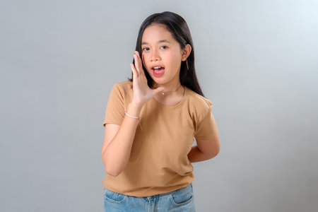 Portrait of a young Asian woman speaking with hand near her mouth, expressing announcement or...
