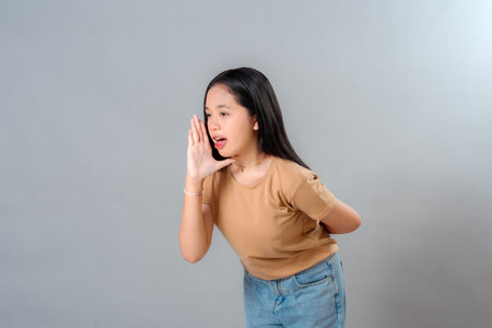 Portrait of a young Asian woman speaking with hand near her mouth, expressing announcement or...