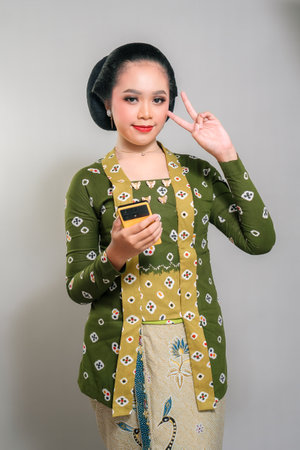Indonesian woman in a green kebaya and batik, holding a smartphone and showing a peace sign with a...