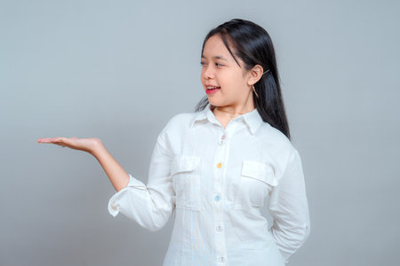 Cheerful Asian girl in white shirt smiling while pointing sideways with open palm, looking at her hand with excited expression, suggesting product placement, isolated on gray background.の写真素材