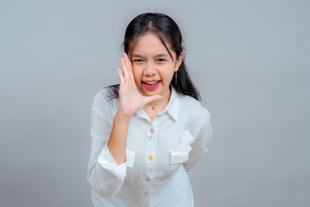 Energetic young Asian woman in white shirt shouting or calling out with her hand beside her mouth, showing a lively and expressive facial expression in a studio portrait.の写真素材