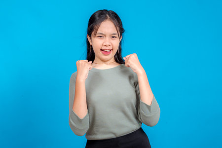 Excited young Asian woman with a confident and determined facial expression, clenching fists in front of her chest. Isolated on blue background. Concept of success and motivation.の写真素材