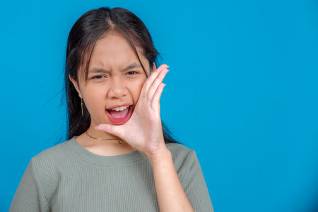 Portrait of a young Asian woman shouting with intense expression, hand cupped near mouth. Her bold, emotional face conveys urgency, protest, or announcement in a striking studio shot.の写真素材