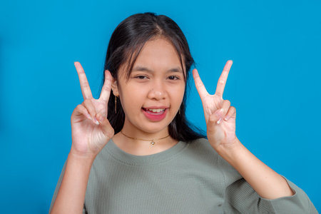 Cheerful young Asian girl showing double peace signs with a playful wink and smile. Confident...