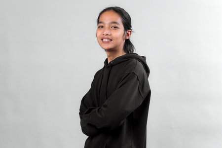 Portrait of a young Asian person with long hair, wearing a black hoodie, smiling confidently with arms crossed, standing against a plain gray background in studio lighting.の写真素材