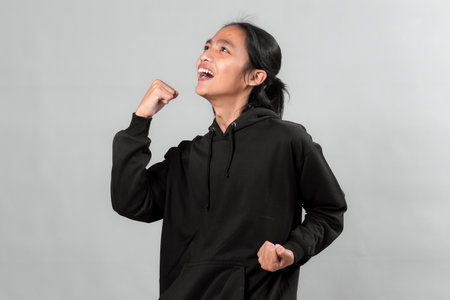 Asian man wearing a black hoodie expressing excitement and success with a triumphant fist gesture,...