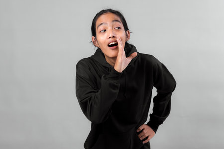 Young Asian woman in black hoodie cupping hand to mouth while speaking or shouting with excited...