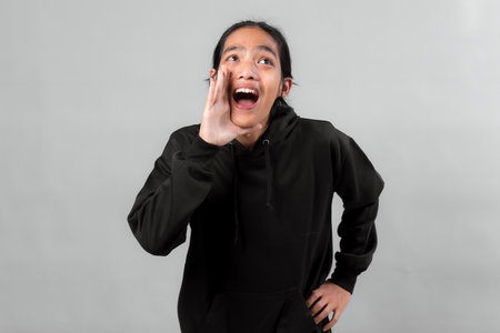 Asian woman wearing a black hoodie shouting with hand near mouth, smiling and looking excited, standing against a plain grey background in a studio setting.の写真素材