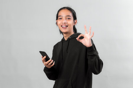 Happy young Asian man in a black hoodie holding a smartphone while showing an OK hand gesture and...
