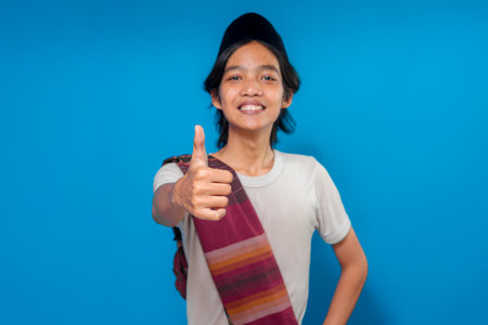 Smiling young Asian man wearing traditional attire giving a thumbs up while standing confidently, expressing approval, satisfaction, and positivity on a blue background.の写真素材