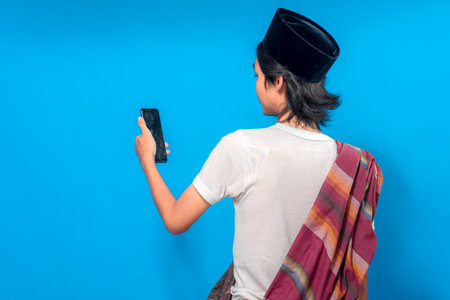 Young Asian man wearing traditional attire seen from behind, looking at his smartphone with a calm...