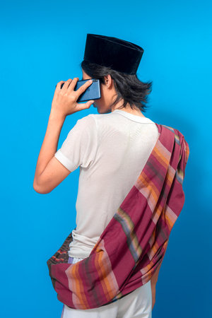 Young man wearing traditional Indonesian attire and peci, seen from behind while talking on a smartphone, showing a serious and attentive expression against a plain blue background.の写真素材