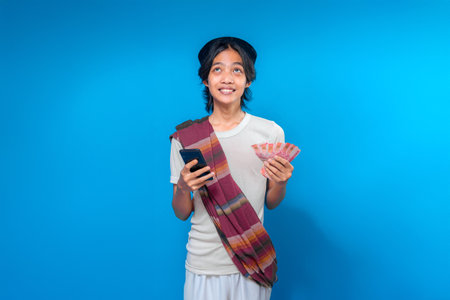 Optimistic Indonesian man wearing peci and traditional sarong over the shoulder, smiling with hopeful eyes while holding smartphone and rupiah cash, standing against blue background.の写真素材