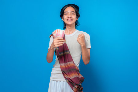 Traditional Indonesian young man in a sarong and peci, winking and smiling cheerfully while holding rupiah money, expressing excitement and happiness on blue background.の写真素材