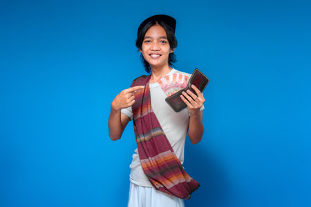 Traditional Indonesian man smiling proudly while pointing at wallet filled with rupiah banknotes. His happy and confident expression reflects joy and financial success.の写真素材