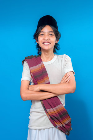 Confident Indonesian young man smiling warmly while crossing arms, wearing traditional sarong and black peci, standing proudly against a solid blue background.の写真素材