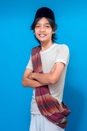 Confident Indonesian young man smiling warmly while crossing arms, wearing traditional sarong and black peci, standing proudly against a solid blue background.の写真素材