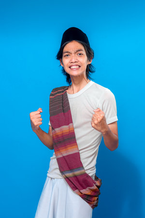 Traditional Indonesian man wearing a black peci and colorful sarong clenches his fists and shows an intense, determined facial expression, symbolizing motivation and strong will.の写真素材