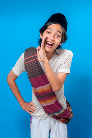 A cheerful young Indonesian man wearing a traditional black peci and colorful sarong over his shoulder, smiling widely and shouting with hand cupped to his mouth.の写真素材