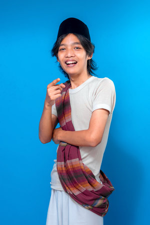 Happy Indonesian man pointing and smiling brightly, wearing traditional sarong and black peci, expressing cheerful and friendly personality on a solid blue background.の写真素材